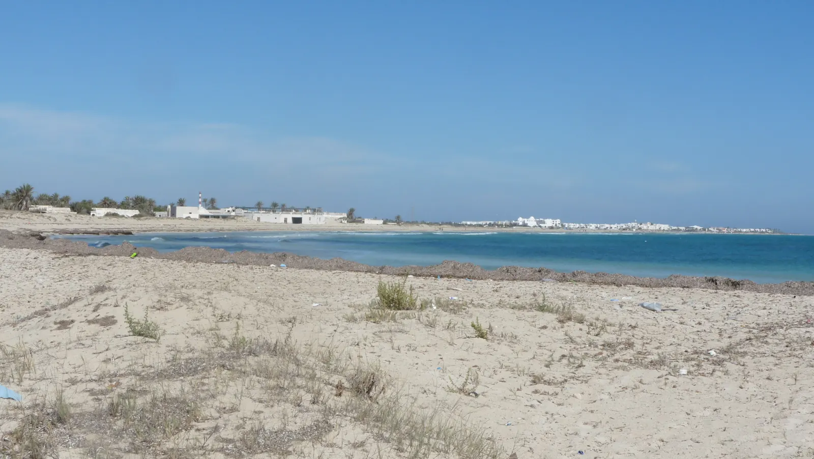 Plage de sable clair presque déserte à Djerba, avec quelques touffes de végétation et une ligne d'hôtels blancs à l'horizon