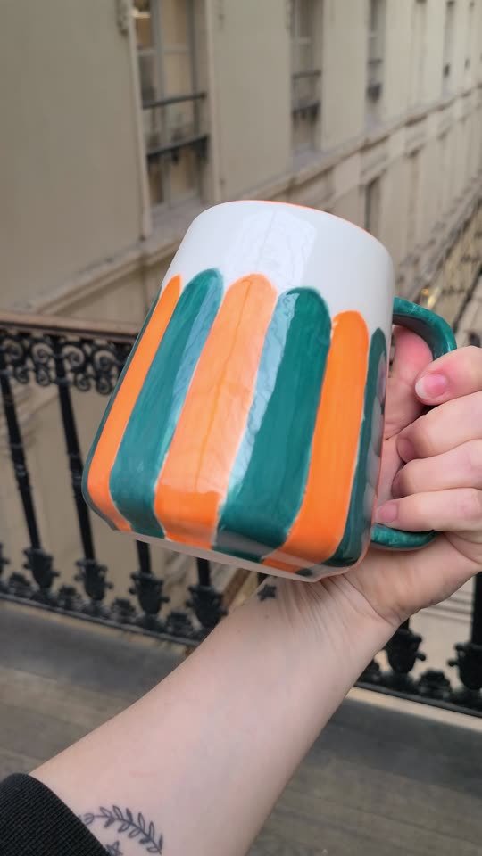 Mug peint de larges rayures orange, vertes et blanches, tenu au-dessus de la balustrade du Passage Pommeraye