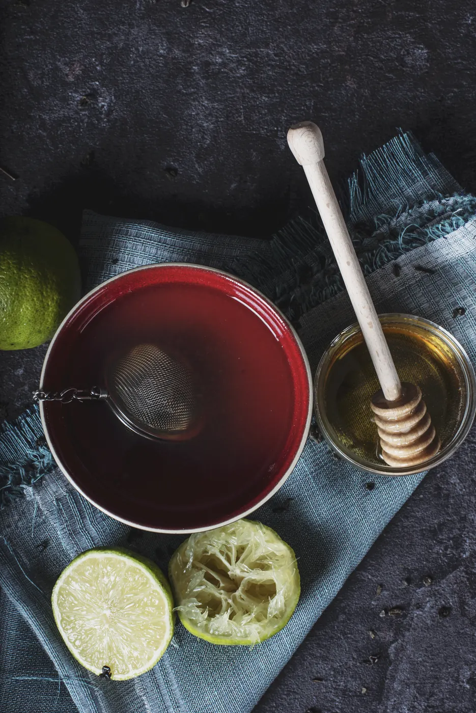 Tasse de tisane rouge avec une boule à thé, un pot de miel et des citrons verts pressés sur un tissu gris