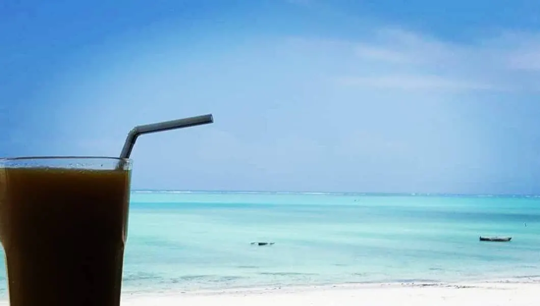 Verre de jus de fruits frais au premier plan devant le lagon turquoise et le sable blanc d'une plage de Zanzibar